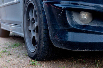 Perspective of front used black car. Can see old style wheels and front bumper with fog lights. Car driving on soil road with green grass at day.