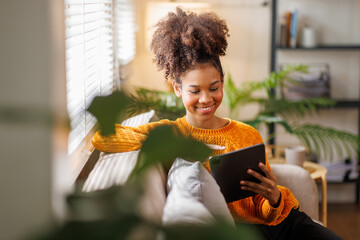 Happy young African American woman using tablet pc on sofa at home. Afro haired woman indoor.