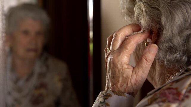 Close up of senior gray haired caucasian woman adjusting her hearing aid kit in front of the mirror. Elderly grandmother enjoying hearing ability after deafness therapy. Deaf patient communication. 