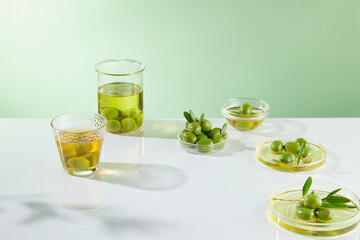 On white table and green background, laboratory glassware full with olive oil, fresh olives and leaves decorated. Blank space in the middle for design product. Advertising photo