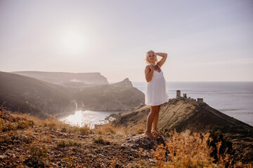 Obraz premium A blonde woman stands on a hill overlooking the ocean. She is wearing a white dress and she is enjoying the view.