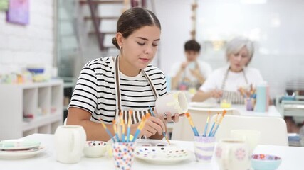 Skilled interested young female artist sitting at table in pottery workshop, painting designs on ceramic cup. Art craft activity concept