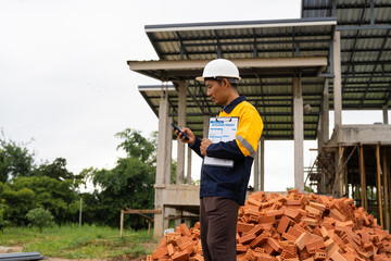 A male architect wearing a hard hat and safety vest checks his laptop while holding a clipboard with house plan papers. He reviews blueprints, construction documents,house structure details on-site.