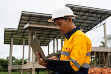 Fototapeta premium A male architect wearing a hard hat and safety vest checks his laptop while holding a clipboard with house plan papers. He reviews blueprints, construction documents,house structure details on-site.
