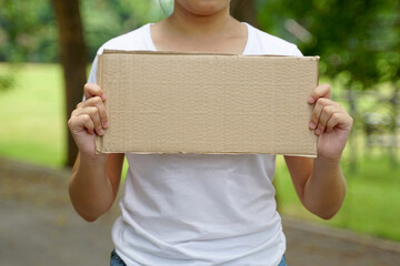 Young Asian woman wearing a white t-shirt stands and holds up a brown cardboard sign with space for...