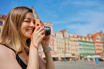 Woman taking photos in picturesque European city square, surrounded by colorful historic buildings. Female tourist captures memories during travel with vintage instant camera