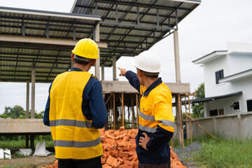 Two male architects wearing hard hats and safety vests discuss construction design on-site. collaborating with the contractor on the house structure, including iron framework, brickwork, and roofing.