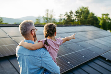 Rear view of dad with girl on roof with solar panels, hugging. Rooftop solar or photovoltaic system. Sustainable future for next generation.