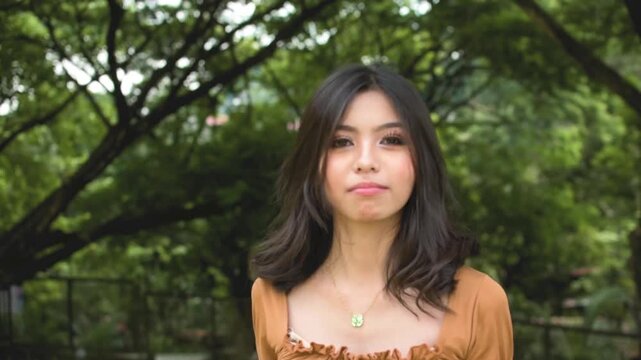 A perky young asian teenager makes heart fingers and other bubbly poses before leaving. Outdoor park scene. Slowed down 50 percent.