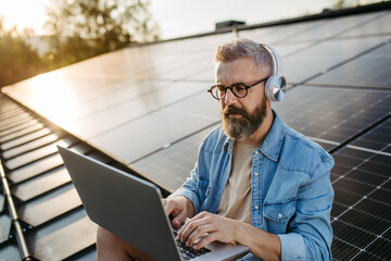 Man sitting on roof full of solar panels, headphones on head, typing on laptop.