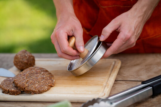 Man in apron preparing meat for hamburgers, standing outdoors. Using burger patty press to shape groud meat.