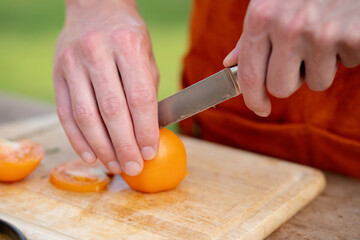 Close up of man holding sharp knife, slicing juicy red tomato. Preparing vegetables for an outdoor barbecue.