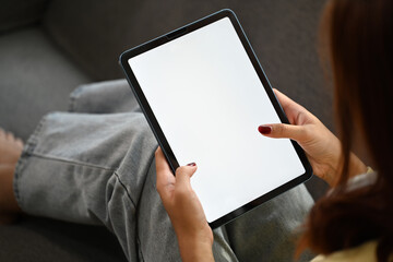 Young woman sitting in cozy sofa holding digital tablet with white blank screen
