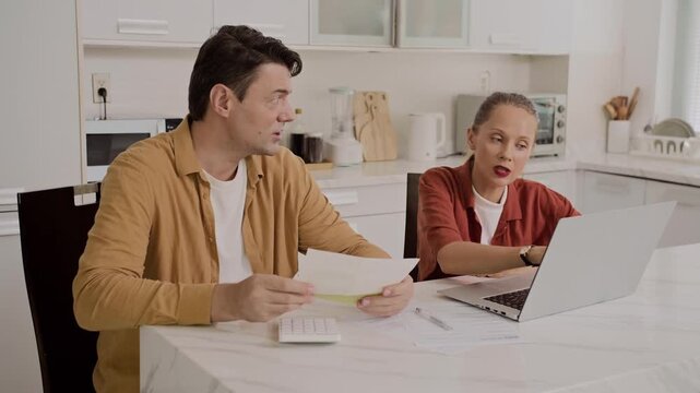 Medium shot of Caucasian couple having arguments while counting bills sitting at kitchen in front of laptop