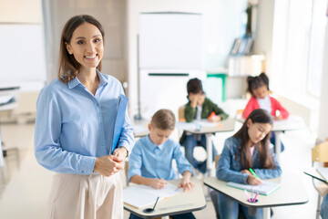 Happy friendly female teacher smiling at camera during lesson at elementary school while pupils writing in copybooks on background in classroom