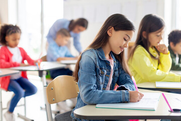 Learning concept. Schoolgirl writing test or taking notes during lesson, sitting at desk in modern classroom interior