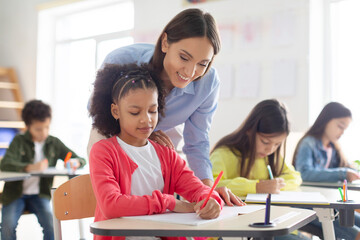 Cheerful female teacher helping girl with homework in classroom, black schoolgirl writing in...