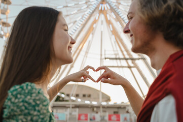 Couple making heart symbol with finger in front of ferris wheel at amusement park