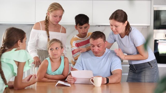  Large family is standing next to confused man sitting near kitchen table. Man reads letter and is upset by bad news from collector, unexpected test results, increase in loan rate