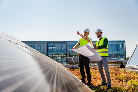 Two technicians inspecting solar panels on the roof of a company building, one pointing to a specific area