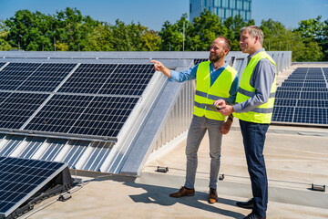 Two workers in safety vests discuss on a rooftop with solar panels while holding a tablet computer