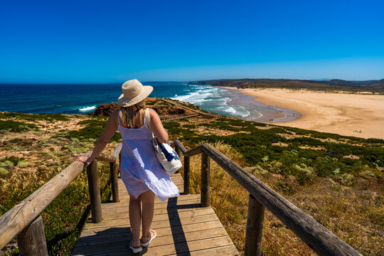 Viewpoint on portuguese beach bordeira praia. Young woman with long blonde hair wearing white dress and hat walking down stairs with beach bag to rocky viewpoint and to sandy beach. Back view