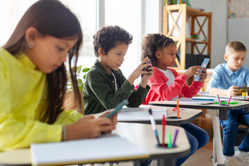 Diverse group of schoolkids using smartphones in classroom, having break during lessons, pupils...