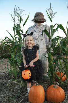 Happy girl holding Jack O' Lantern in front of scarecrow near pumpkin patch