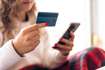 Unrecognizable young woman using smartphone and credit card while sitting at home with laptop at table