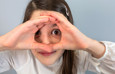 A young girl with a playful expression looks through a binocular shape made with her hands against a gray background