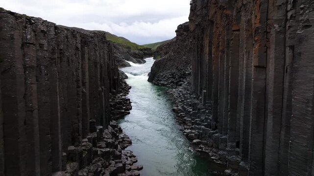 A drone captures the breathtaking Stu&eth;lagil canyon in Iceland, flying between towering rock formations above the river. This aerial view highlights the canyon's dramatic and rugged geological features