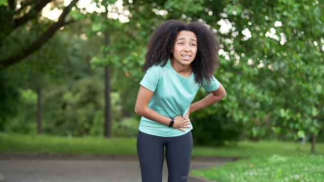 Young sporty african american female suffering from stomach pain during morning run in urban city park. Upset black woman has poisoning, spasms, menstruation, constipation or digestive problems