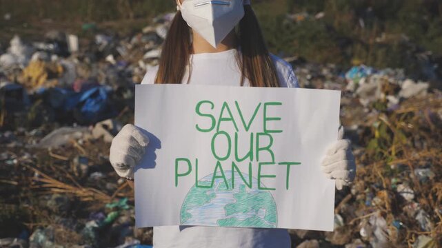 Small girl holds in hands placard of environmental movement for saving earth. Little eco volunteer in protective mask advocating for nature conservation against background of dirty dump. Crane shot