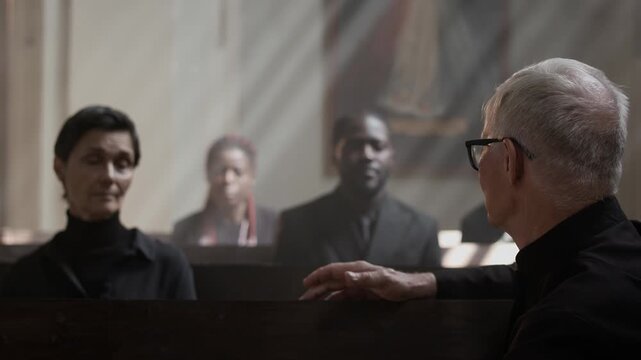 Senior pastor sitting sideways on wooden pew in catholic church and talking to parishioners after funeral service