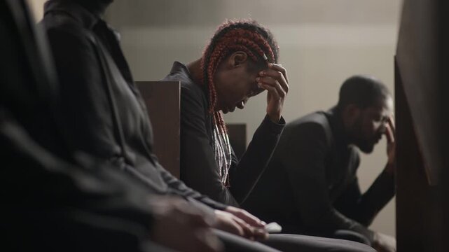Rack focus shot of two unrecognisable people and two grieving African American friends sitting on wooden pew with their heads down at funeral service