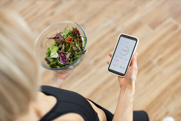 Woman in fitness clothing tracking daily calorie intake on smartphone while holding bowl of fresh...