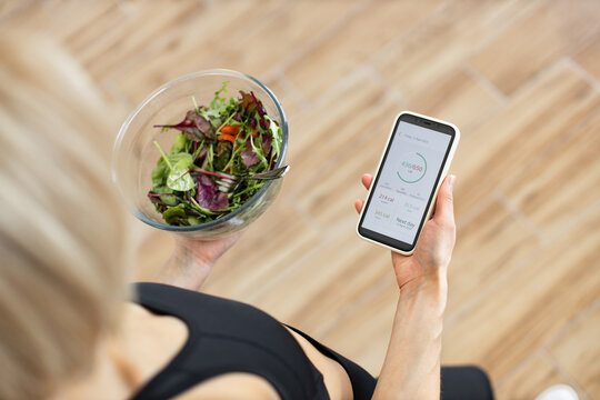 Woman in fitness clothing tracking daily calorie intake on smartphone while holding bowl of fresh salad. Concept of healthy eating, fitness, nutrition, and technology.