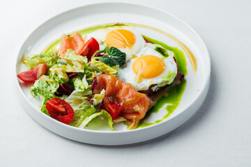 Breakfast, two eggs served on toasted bread with a green herb sauce. Accompanied fresh salad, cherry tomatoes, and smoked salmon slices, all elegantly presented on a white plate and white background