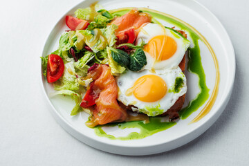 Breakfast, two eggs served on toasted bread with a green herb sauce. Accompanied fresh salad, cherry tomatoes, and smoked salmon slices, all elegantly presented on a white plate and white background