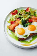 Breakfast, two eggs served on toasted bread with a green herb sauce. Accompanied fresh salad, cherry tomatoes, and smoked salmon slices, all elegantly presented on a white plate and white background