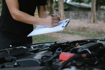 A male motor mechanic stands in a workshop, performing a safety inspection on a car engine. Surrounded by tools and equipment, he diagnoses and repairs vehicle issues.