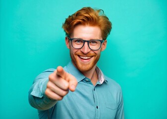 Attractive reddish-haired male in glasses and casual attire, pointing at the viewer with a bright smile, against a vibrant turquoise background, conveying a sense of approachability.