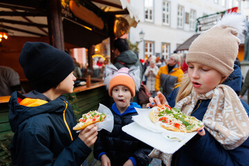 Three cute children kids wear warm winter clothing enjoy freshly made street food langos bread at german Christmas outdoor market  winter festival. Festive atmosphere holiday spirit concept