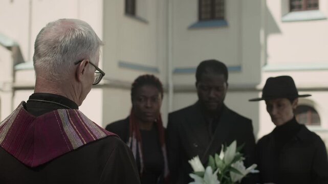 Back shot of Caucasian senior pastor reading prayer in front of three family members at outdoor funeral service