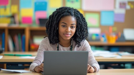 A young teacher of African American descent sitting at her desk in a modern classroom, working on a laptop, preparing lesson plans, and utilizing digital resources to enhance her teaching methods