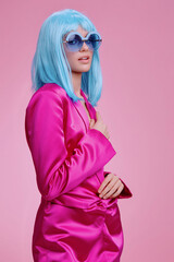 Woman in a pink suit, with blue hair and blue sunglasses, standing against a pink background