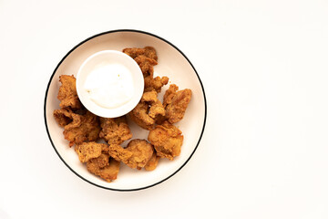 crispy, golden-brown pieces of fried chicken, most likely popcorn chicken or similar bite-sized chicken nuggets, arranged around a small white bowl of dipping sauce on a white plate with a dark rim. A