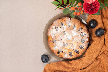 Sweet plum pie with sugar powder on beige table with orange tablecloth, plum fruits and flowers, top view, empty copy space. Seasonal fall traditional dessert, homemade cake