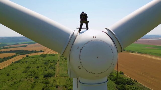 Aerial high angle footage of industrial rope access technician working on a wind turbine generator. Drone shot of skilled professional workers working at high altitude doing repairs of damaged blade.

