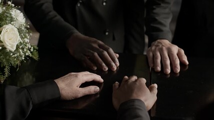 Close up view of hands of four unrecognisable people in black suits keeping them on coffin lid during indoor funeral ceremony
